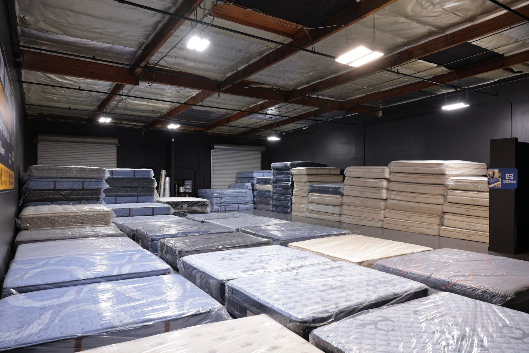 The warehouse floor at John's Mattress Liquidation, rows of plastic-wrapped mattresses laid out with more stacked along the back wall